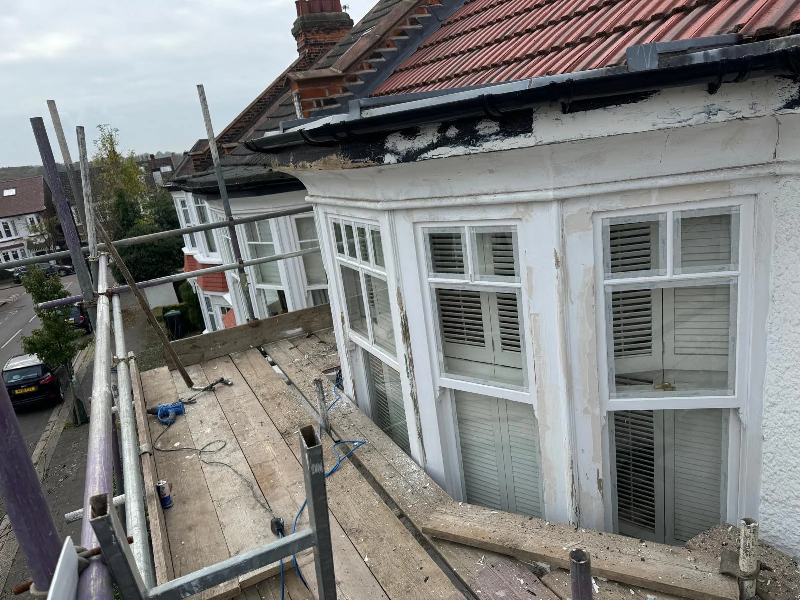 Bay window elevation with peeling paint and stripped fascia viewed from scaffold before restoration